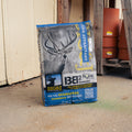 A bag of Big & J® BB2 Enhance Corn Pellet Attractant deer attractant stands on a concrete floor in front of a weathered wooden door, with stacked terracotta pots visible in the background.