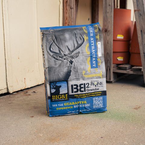A bag of Big & J® BB2 Enhance Corn Pellet Attractant deer attractant stands on a concrete floor in front of a weathered wooden door, with stacked terracotta pots visible in the background.