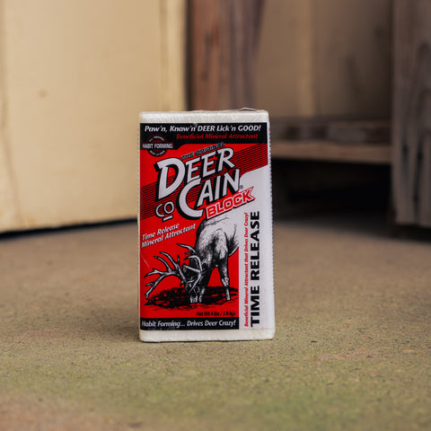 A single box of Deer Cain Block time-release natural attractant standing upright on a textured ground with a blurred wood background.