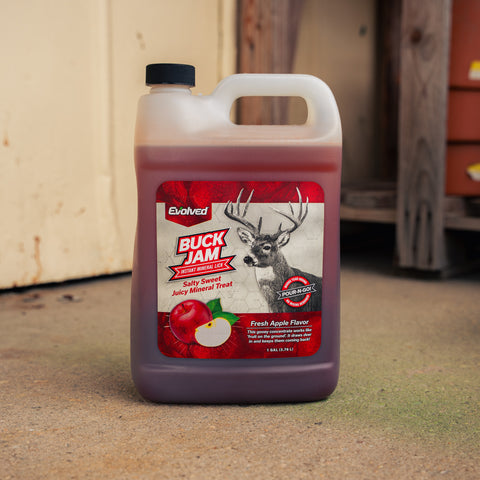 A 5-gallon plastic jug of Evolved Buck Jam salty sweet mineral treat in fresh apple flavor, filled with a dark red liquid, set on a concrete floor in front of a rustic wooden door.