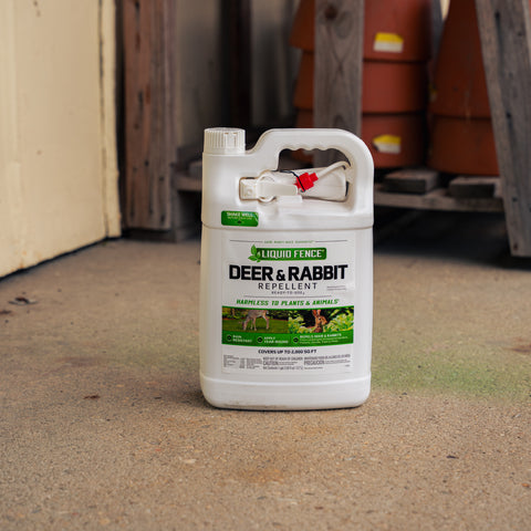 A white plastic bottle of Liquid Fence Deer & Rabbit Repellent standing on a concrete floor in a warehouse or shed setting.