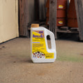 A white plastic bottle of Repels-All Animal Repellent with a yellow label and tan cap, standing on a concrete floor in a rustic workshop or barn setting.