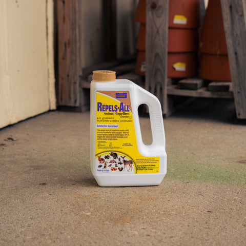 A white plastic bottle of Repels-All Animal Repellent with a yellow label and tan cap, standing on a concrete floor in a rustic workshop or barn setting.