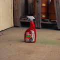 A red spray bottle of Deer Stopper stands on a concrete floor in a workshop or garage setting with wooden shelves and terracotta pots in the background.