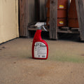 A red spray bottle of Deer Stopper stands on a concrete floor in a workshop or garage setting with wooden shelves and terracotta pots in the background.