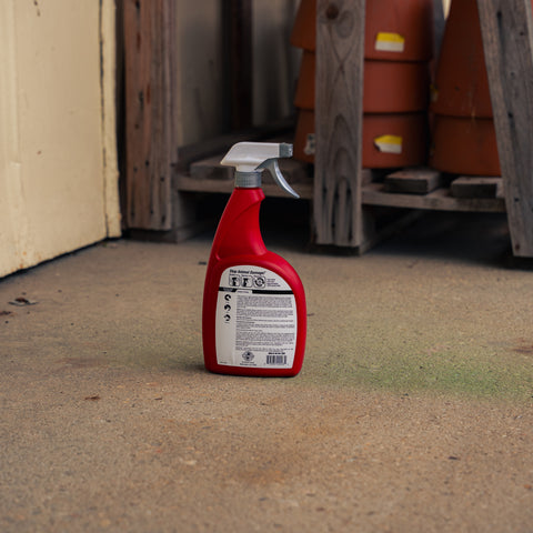 A red spray bottle of Deer Stopper stands on a concrete floor in a workshop or garage setting with wooden shelves and terracotta pots in the background.