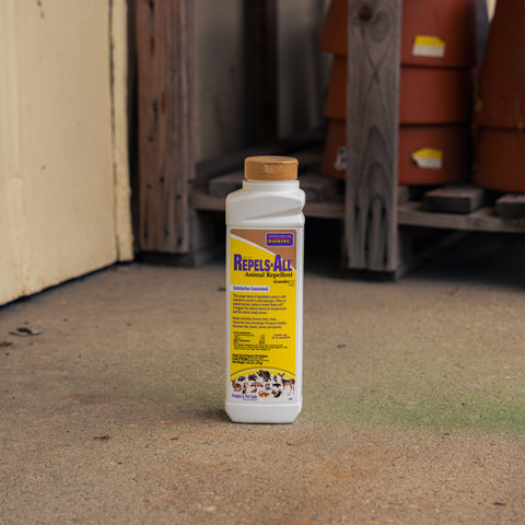 A bottle of Repels-All Animal Repellent standing on a concrete floor in an outdoor or semi-outdoor storage area with wooden pallets and terracotta pots in the background.