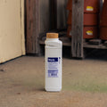 A bottle of Repels-All Animal Repellent standing on a concrete floor in an outdoor or semi-outdoor storage area with wooden pallets and terracotta pots in the background.