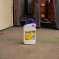 A white plastic bottle of Repels-All with a translucent purple cap, standing on a concrete floor in a workshop or garage setting.