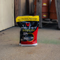 A bag of Victor Snake-A-Way snake repelling crystals standing on a concrete floor in an outdoor or industrial setting with wooden posts and terracotta barrels in the background.