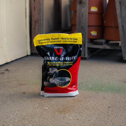 A bag of Victor Snake-A-Way snake repelling crystals standing on a concrete floor in an outdoor or industrial setting with wooden posts and terracotta barrels in the background.