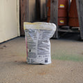 A white plastic bag of snake repellent stands upright on a concrete floor in a warehouse or storage area, with wooden pallets and terracotta pots blurred in the background.
