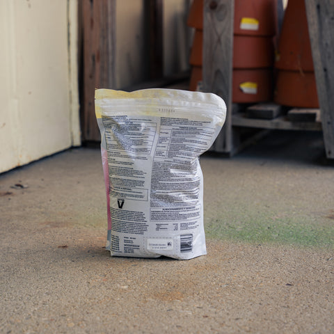 A white plastic bag of snake repellent stands upright on a concrete floor in a warehouse or storage area, with wooden pallets and terracotta pots blurred in the background.