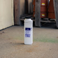 A bottle of Mole & Gopher Killer standing on a concrete floor in a garage or shed with wooden pallets and stacked terracotta pots in the background.