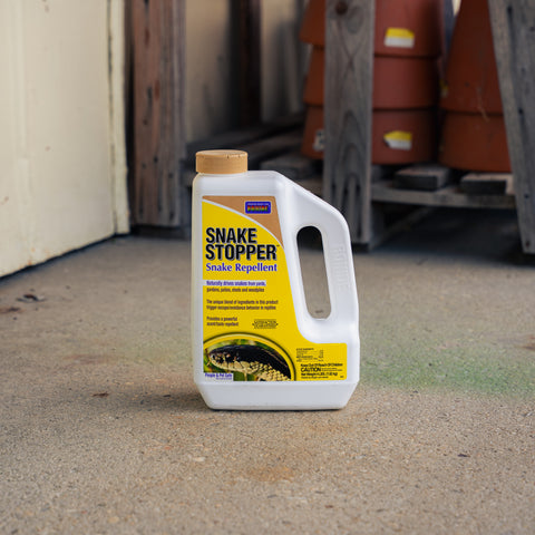 A white plastic bottle of Snake Stopper Snake Repellent with a yellow label and tan cap, standing on a concrete floor in a warehouse or shed setting.