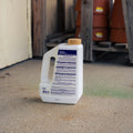 A white plastic bottle of Snake Stopper Snake Repellent with a yellow label and tan cap, standing on a concrete floor in a warehouse or shed setting.