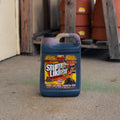 A large plastic jug of Stump Likker stands on a concrete floor in front of a wooden shed with stacked terracotta pots in the background.