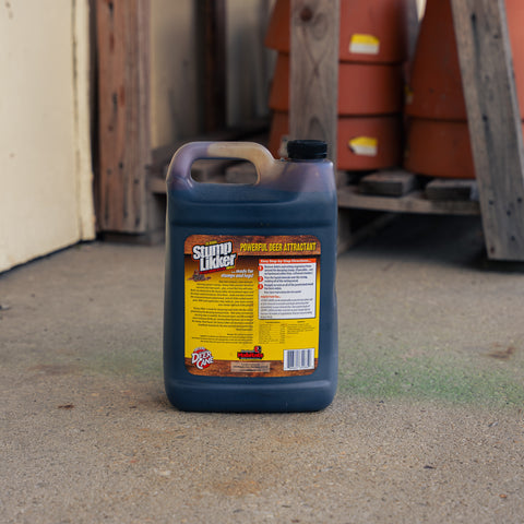 A large plastic jug of Stump Likker stands on a concrete floor in an outdoor or workshop setting with wooden posts and terracotta pots in the background.