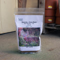 A 4 lb bag of 'Mill Perennial - Clover Blend' deer plot mix stands on a concrete floor in an outdoor shed setting. The clear plastic bag features a white paper label with a photo of a deer and the brand name 'The Mill'.
