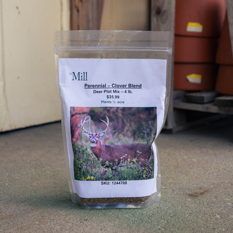 A 4 lb bag of 'Mill Perennial - Clover Blend' deer plot mix stands on a concrete floor in an outdoor shed setting. The clear plastic bag features a white paper label with a photo of a deer and the brand name 'The Mill'.