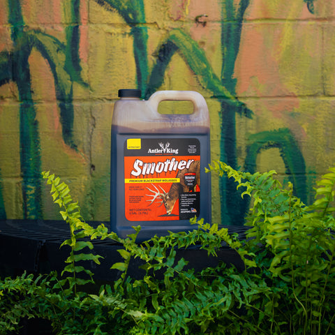 A bottle of Antler King Smoother deer molasses solution, placed on a black surface with green ferns in the foreground and a painted brick wall in the background.