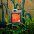 A bottle of Antler King Smoother deer molasses solution, placed on a black surface with green ferns in the foreground and a painted brick wall in the background.