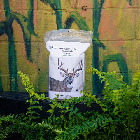 A bag of Mill Deer Plot Mix - 6 lb. Annual Mix plant % acre product standing on a black surface with green ferns in the foreground and a painted brick wall behind it.