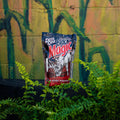 A bag of Deer Cane Black Magic deer attractant standing upright on a black surface, surrounded by green ferns in front of a graffiti-covered wall.