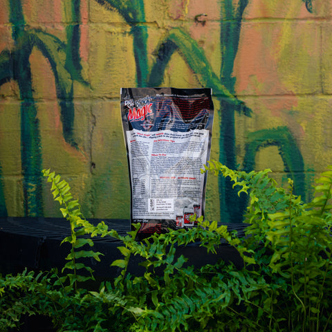 A bag of Deer Cane Black Magic deer attractant standing upright on a black surface, surrounded by green ferns in front of a graffiti-covered wall.