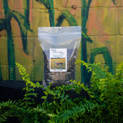 A clear stand-up pouch of Mill #5 Sunn Hemp, filled with dark granular fertilizer, displayed on a black shelf in front of a green and orange painted brick wall with live plants.