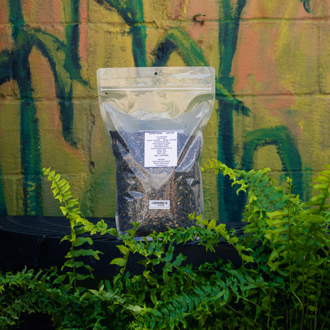 A clear stand-up pouch of Mill #5 Sunn Hemp, filled with dark granular fertilizer, displayed on a black shelf in front of a green and orange painted brick wall with live plants.