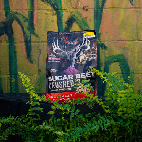 A large bag of Wildgame Sugar Beet Crushed Pellet Feed stands upright outdoors in front of a graffiti-covered wall, surrounded by green plants.