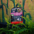 A large bag of Wildgame Sugar Beet Crushed Pellet Feed stands upright outdoors in front of a graffiti-covered wall, surrounded by green plants.