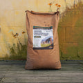 A large brown paper bag of Buck Stampede deer plot mix stands on a wooden deck against a textured yellow and green painted wall.