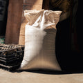 A large off-white woven burlap sack standing upright on a concrete floor in a warehouse or storage area, with wooden beams, black pallets, and a dark doorway in the background.