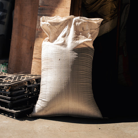 A large off-white woven burlap sack standing upright on a concrete floor in a warehouse or storage area, with wooden beams, black pallets, and a dark doorway in the background.