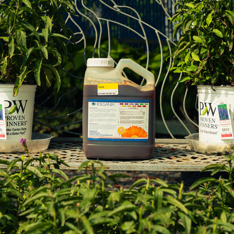 A large plastic jug of HGX PRO Bio 800 Tree and Shrub biostimulant sits on a metal garden table outdoors, flanked by potted plants on either side.