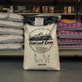 A 50 lb bag of Cracked Corn standing on a store shelf, surrounded by other bags of pet food. The bag is off-white with black graphics and text, featuring an illustration of a barn, corn, and two chickens.
