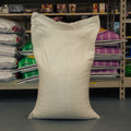 A large off-white woven bulk sack standing upright on a gray floor in front of metal shelving stocked with other bags of materials.