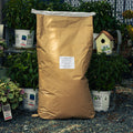 A large brown paper bag of Sunflower Medium Chips stands upright on gravel outdoors, surrounded by potted green plants and a small birdhouse. A white label is printed on the front of the bag.