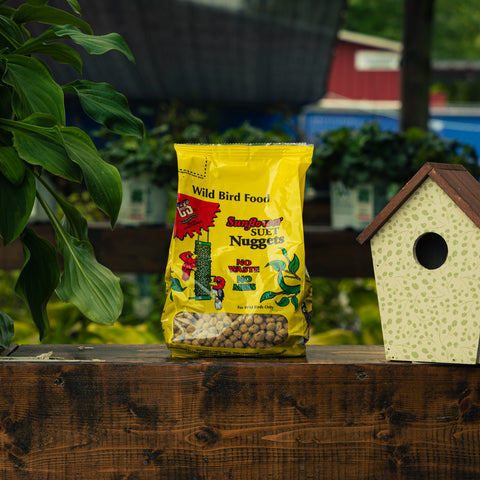 A bag of Sunflower Suet Nuggets wild bird food sits on a wooden ledge next to a decorative birdhouse, with green foliage and a garden background.