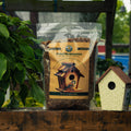 A bag of Dried Mealworms by Bird Pro is displayed on a wooden surface next to a decorative birdhouse, with a garden background.