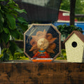 A decorative octagonal sun figurine made from sunflower seeds, packaged in clear plastic wrap and displayed on a wooden surface next to a small birdhouse, with green plants and an outdoor background.