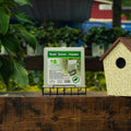 A package of Suet Saver Feeder displayed on a small black metal stand on a wooden surface, with a birdhouse to the right and green foliage in the foreground, set against a blurred outdoor background.