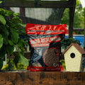 A bag of Wild Bird Food Dried Mealworm High Protein bird feed placed on a wooden table outdoors, next to a small birdhouse and green plants.