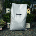 A large white plastic bag of Dried Mealworms standing upright on gravel, surrounded by potted green plants and a small birdhouse in a garden setting.