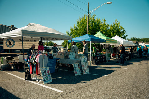 Outdoor farmers market scene with several canopy tents set up on a paved street. A white tent in the foreground displays clothing racks and signs, with people standing and browsing nearby.