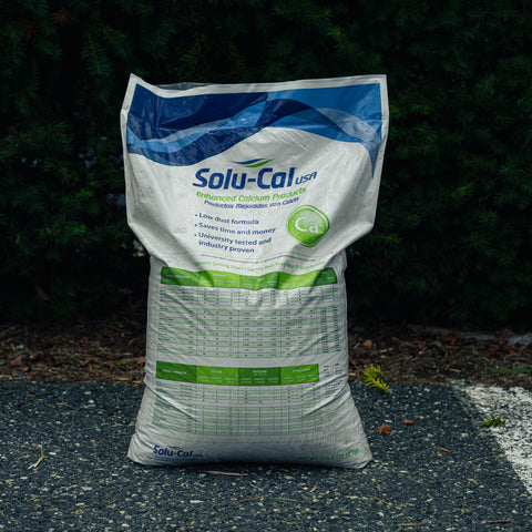 A large white bag of Solu-Cal USA Enriched Cactus Products Potassium Fertilizer stands on asphalt in front of a dark green hedge. The bag features blue and green branding and text, including a prominent 'Ca' circular badge.