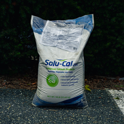 A large white bag of Solu-Cal USA Enhanced Calcium Products standing on asphalt outdoors with dark green foliage in the background.