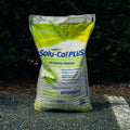 A large bag of Solu-Cal Plus Enhanced Calcitic Lime fertilizer, featuring a white and green design with blue text, standing on a paved surface with dark green foliage in the background.